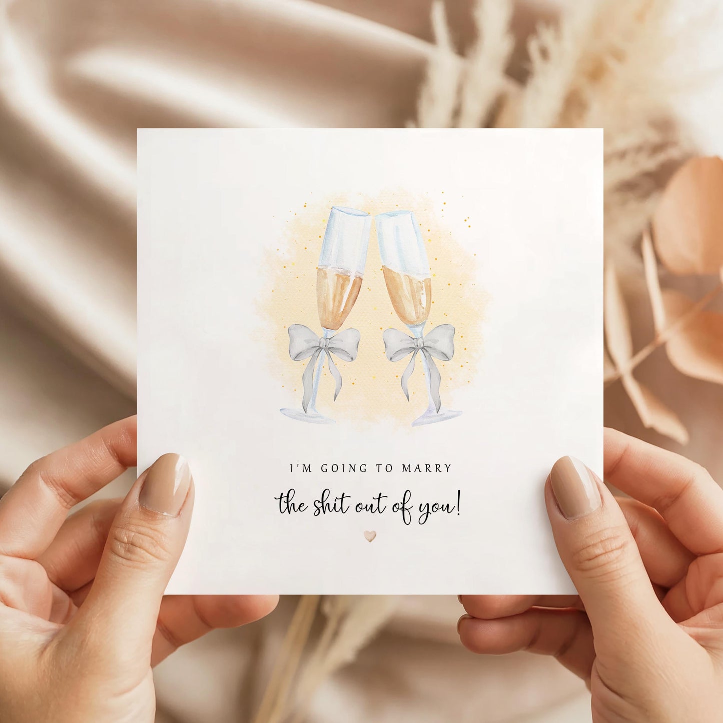 Funny wedding day card with watercolour champagne flutes tied with grey bows and gold confetti, with the text “I’m going to marry the shit out of you!” on a white square card, photographed held in hands against a soft neutral beige background.