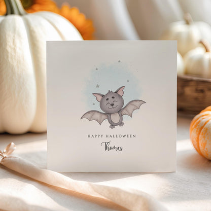 Personalised Halloween card featuring a cute smiling watercolour bat with a custom name underneath and the words "Happy Halloween" on a white greeting card.