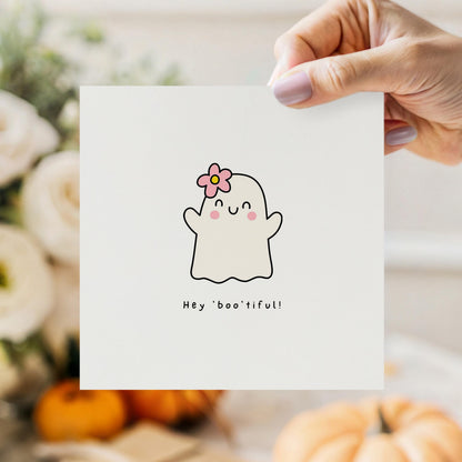 Cute Halloween greeting card featuring a smiling ghost with a pink flower in her hair and the playful message "Hey Boo'tiful!"