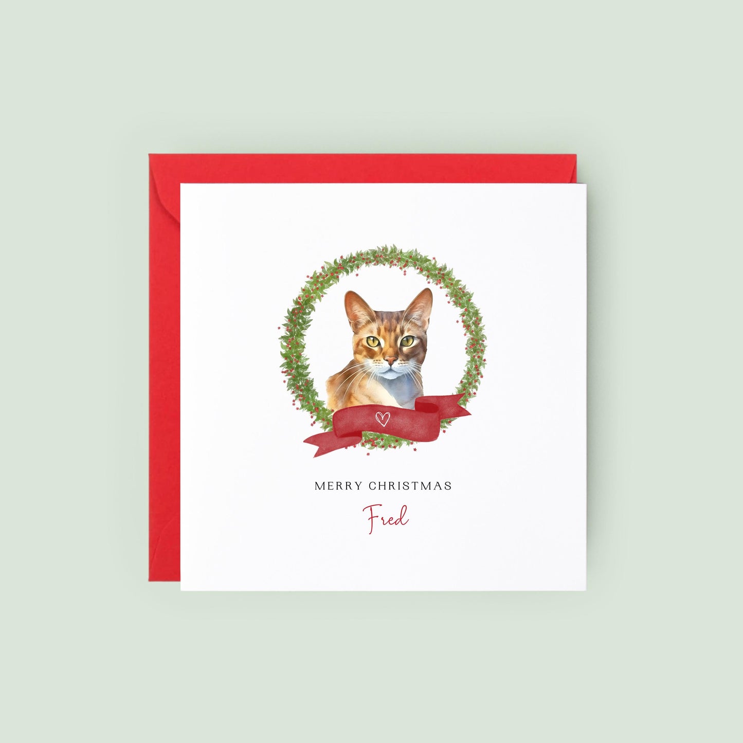 Personalised Abyssinian cat Christmas card featuring a watercolour cat illustration inside a festive wreath with a red ribbon, text reads “Merry Christmas Fred”.