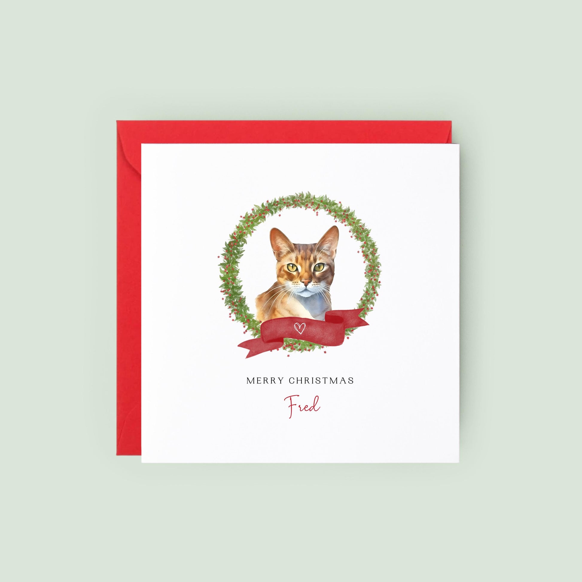 Personalised Abyssinian cat Christmas card featuring a watercolour cat illustration inside a festive wreath with a red ribbon, text reads “Merry Christmas Fred”.