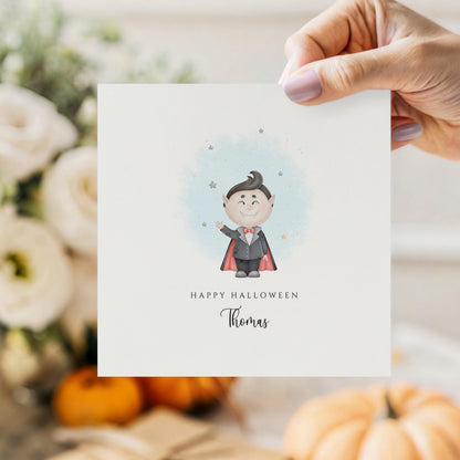 Personalised Halloween card featuring a cute smiling vampire in a cape with a custom name underneath and the words "Happy Halloween" on a white greeting card.