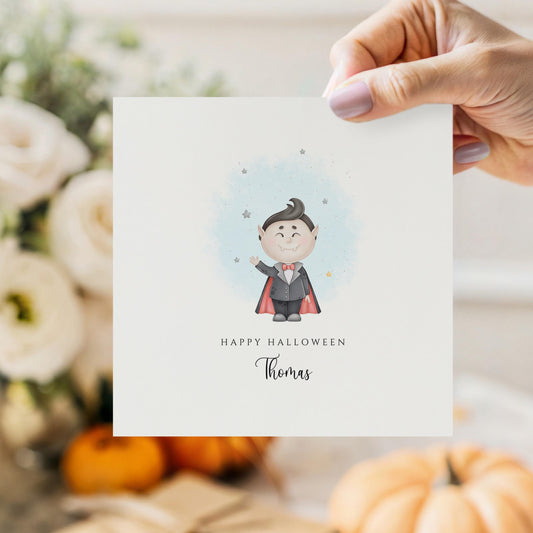 Personalised Halloween card featuring a cute smiling vampire in a cape with a custom name underneath and the words "Happy Halloween" on a white greeting card.