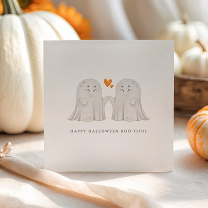 Cute Halloween greeting card featuring two smiling ghost characters high-fiving beneath orange hearts with the message "Happy Halloween Boo'tiful."