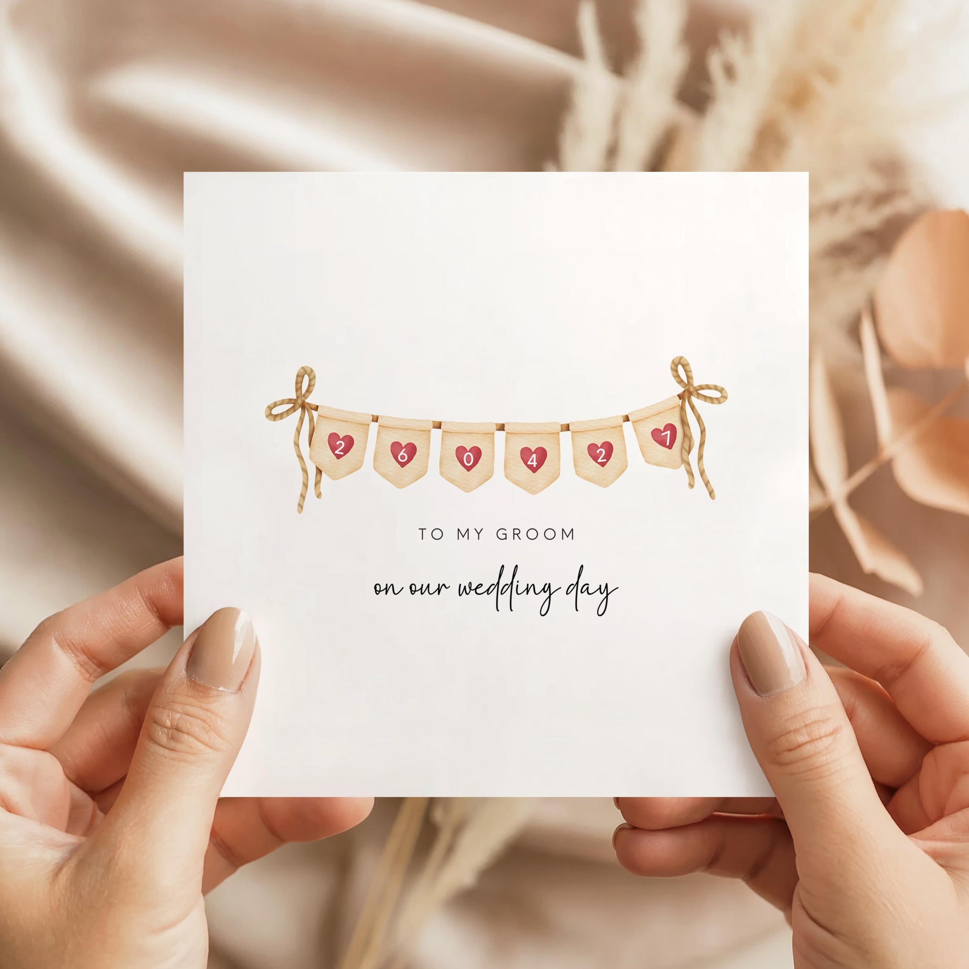 Hands holding a square wedding card with heart bunting showing a personalised date and initials, text saying to my groom on our wedding day, soft neutral background.