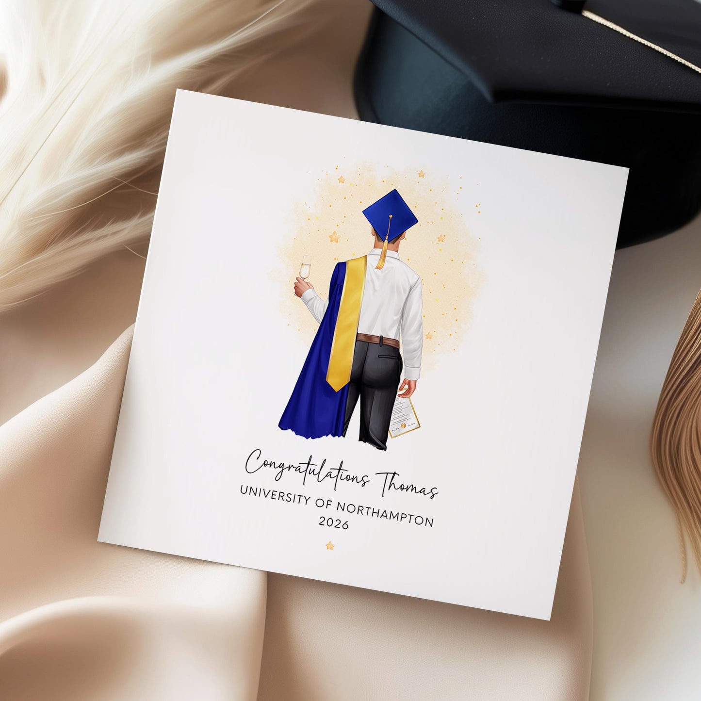 Personalised graduation card for him featuring a male graduate in blue and gold academic robes with custom name, university and graduation year.